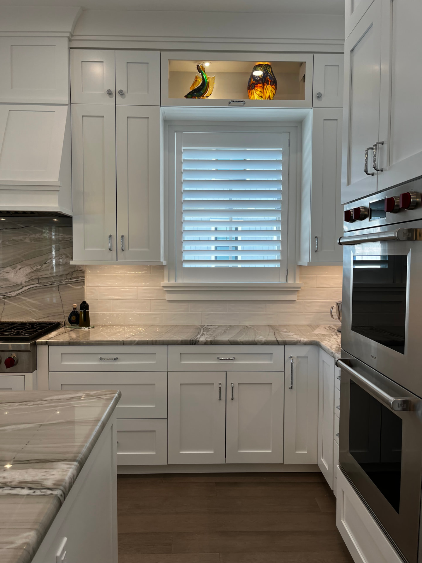 Shaker wall cabinets framing a shuttered window with a lit display niche above, beside a double wall oven