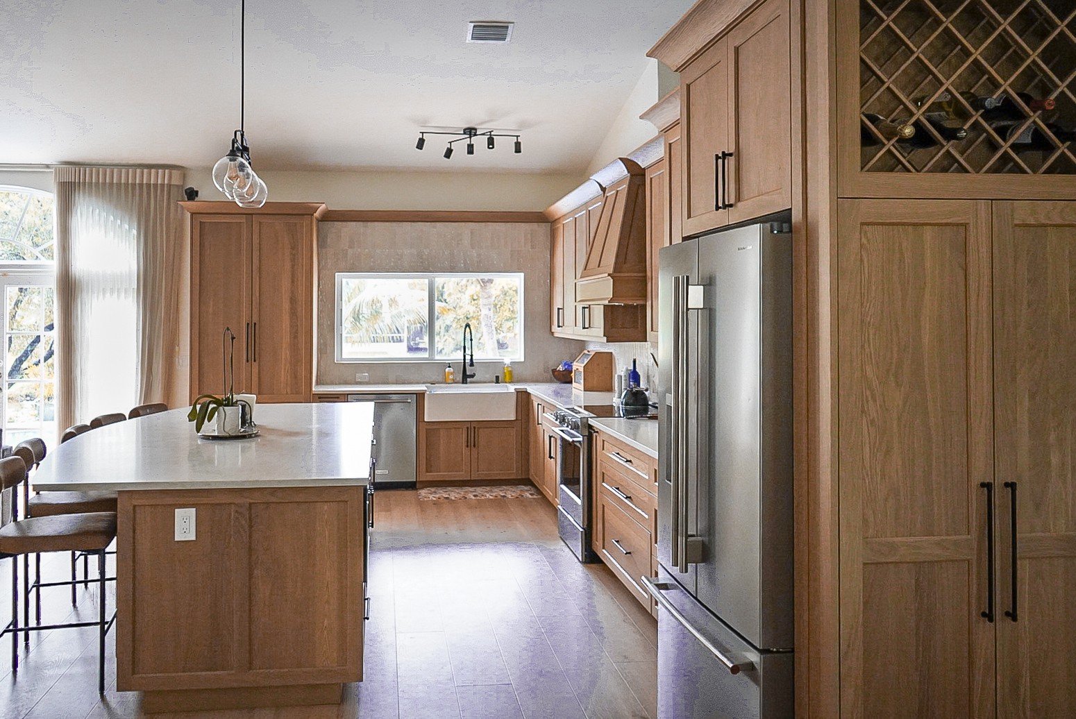 Wide view of the white oak kitchen showing the island, farmhouse sink, paneled refrigerator surround, and wine lattice cabinet