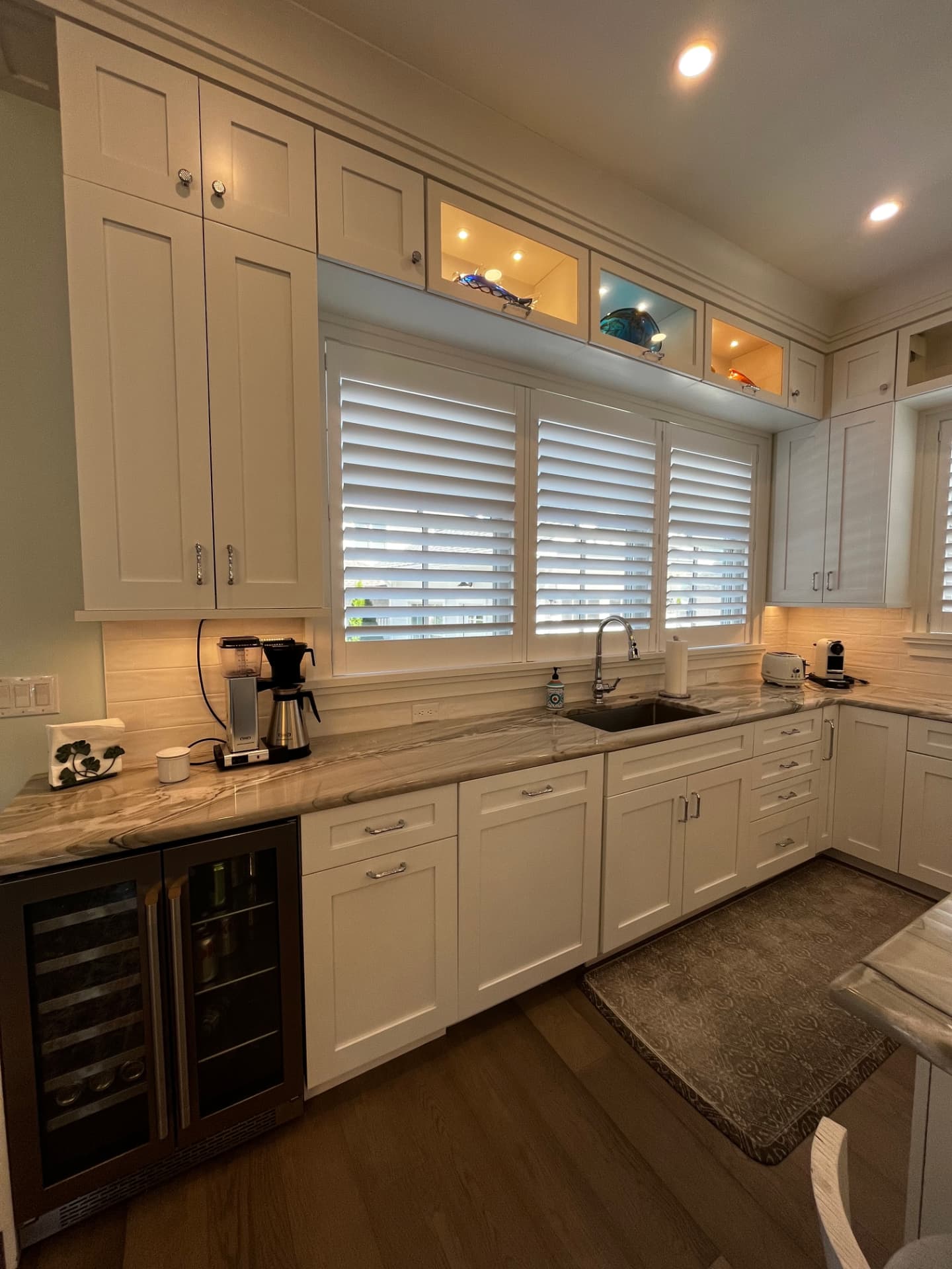 Sink wall with lit glass display cabinets above the windows, white shaker base cabinets, and a built-in beverage cooler