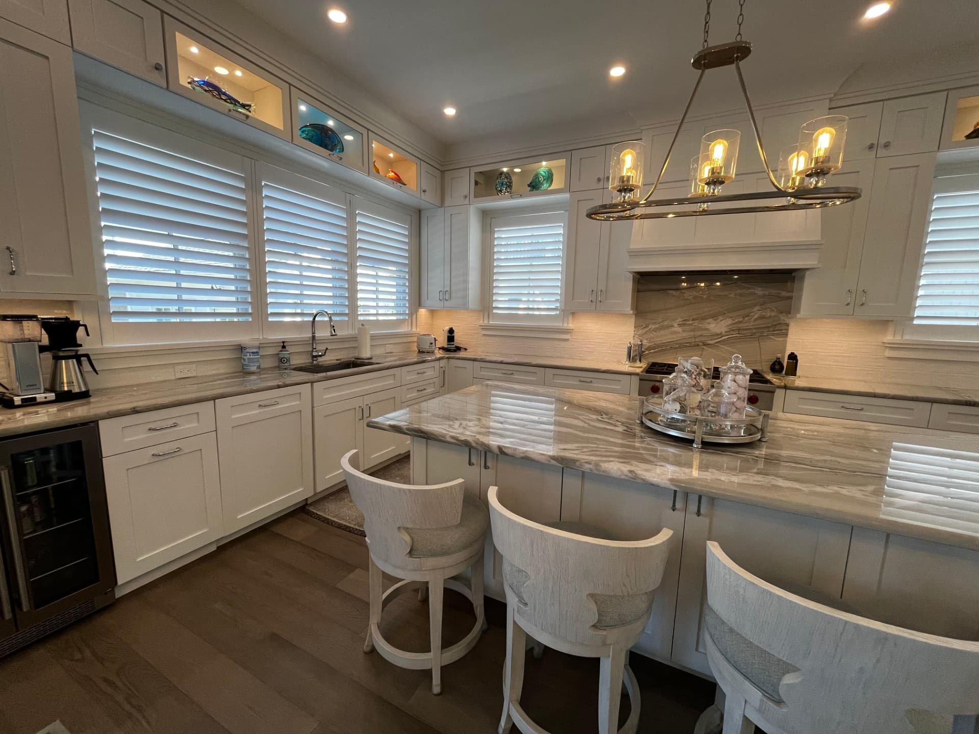 White shaker kitchen with lit glass display niches above the windows, quartzite island, and beverage cooler