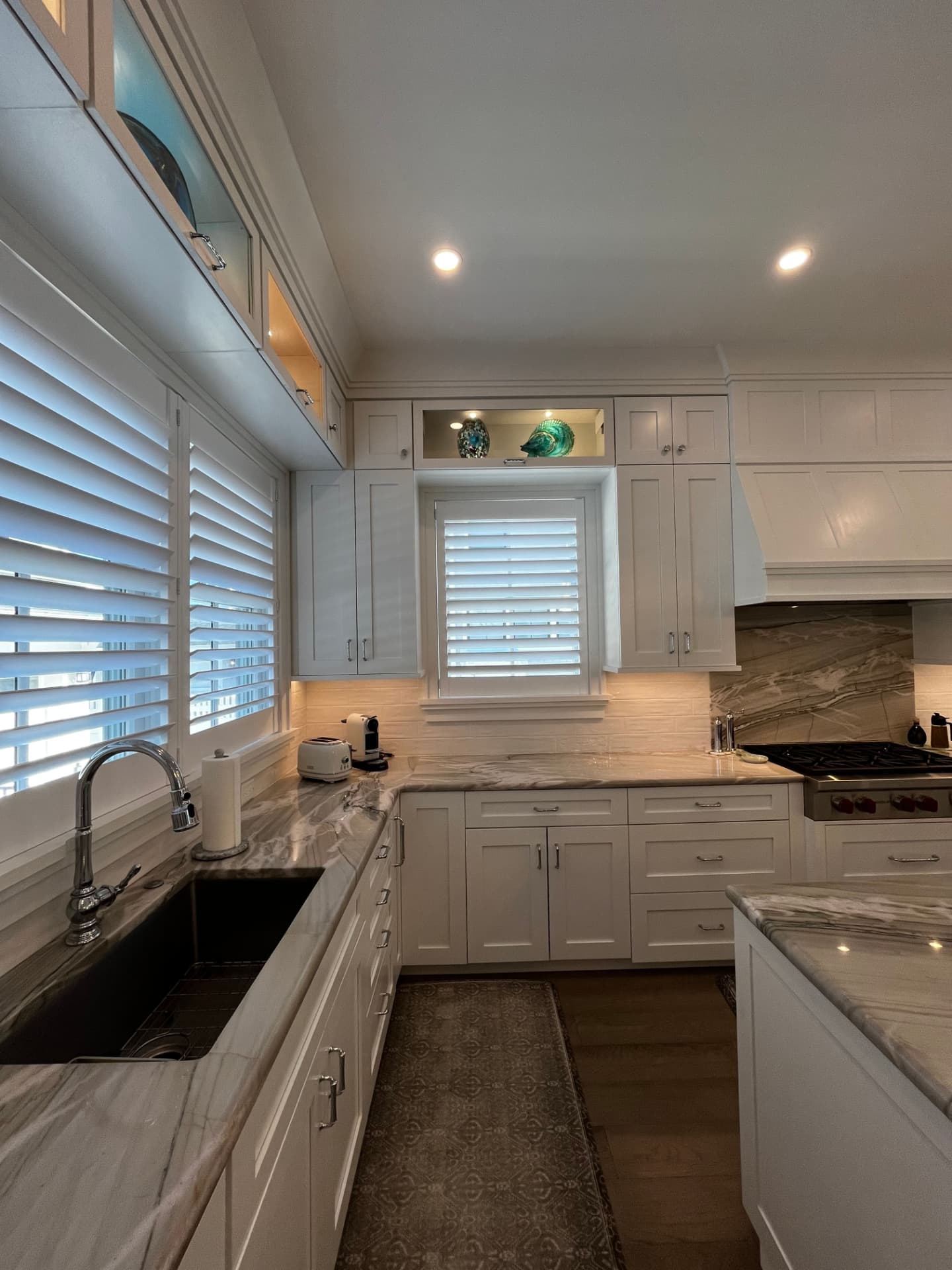 Corner view of white shaker cabinetry with lit glass display cabinets above the sink window and quartzite countertops