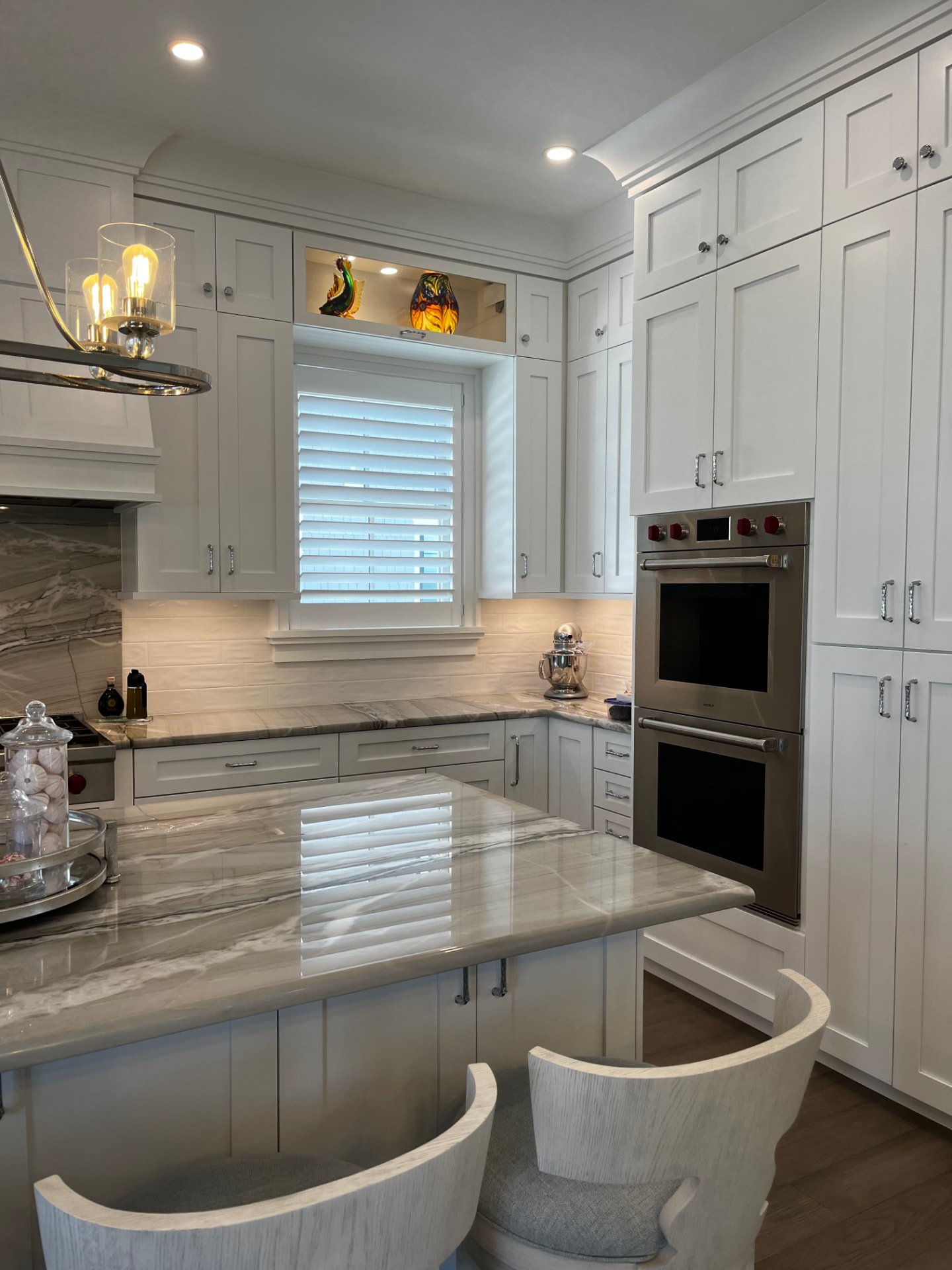 Tall white shaker cabinetry beside a double wall oven, with a lit display niche above the window and a quartzite island counter