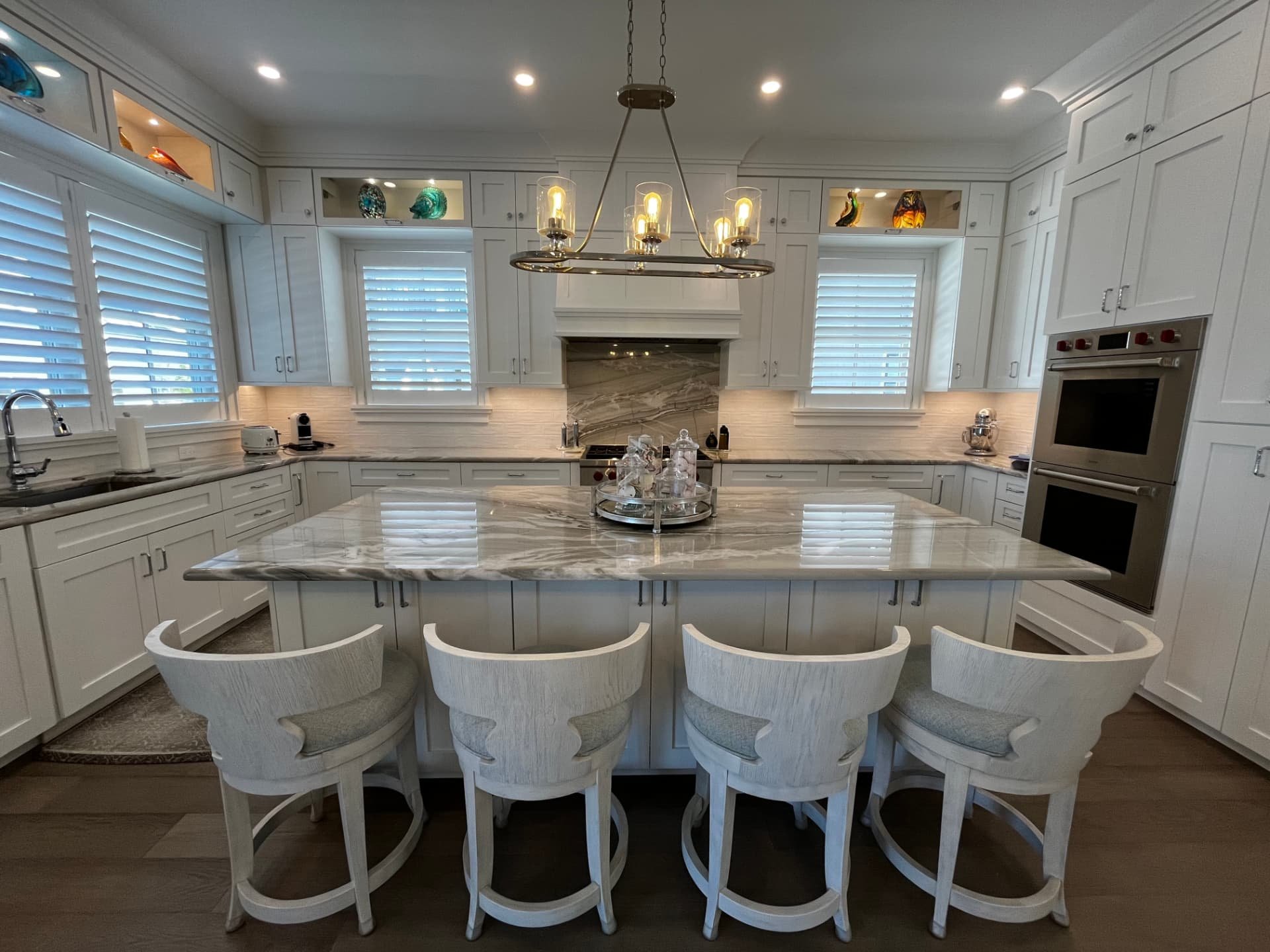 White shaker kitchen with a large quartzite island, four white counter stools, glass-front lit display cabinets, and a custom hood