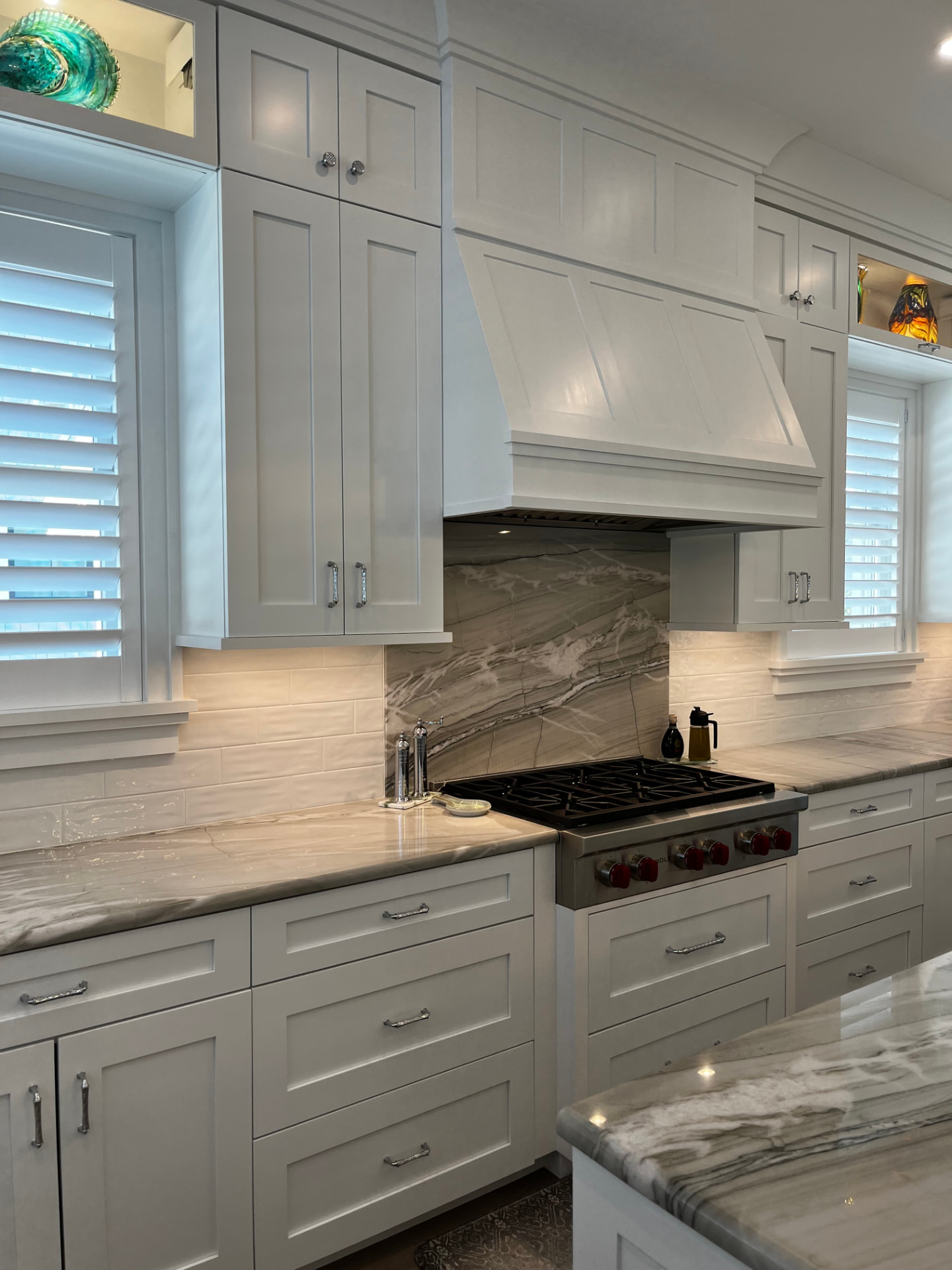 Custom white hood flanked by tall shaker cabinets, with a quartzite slab backsplash and lit display niche above the window
