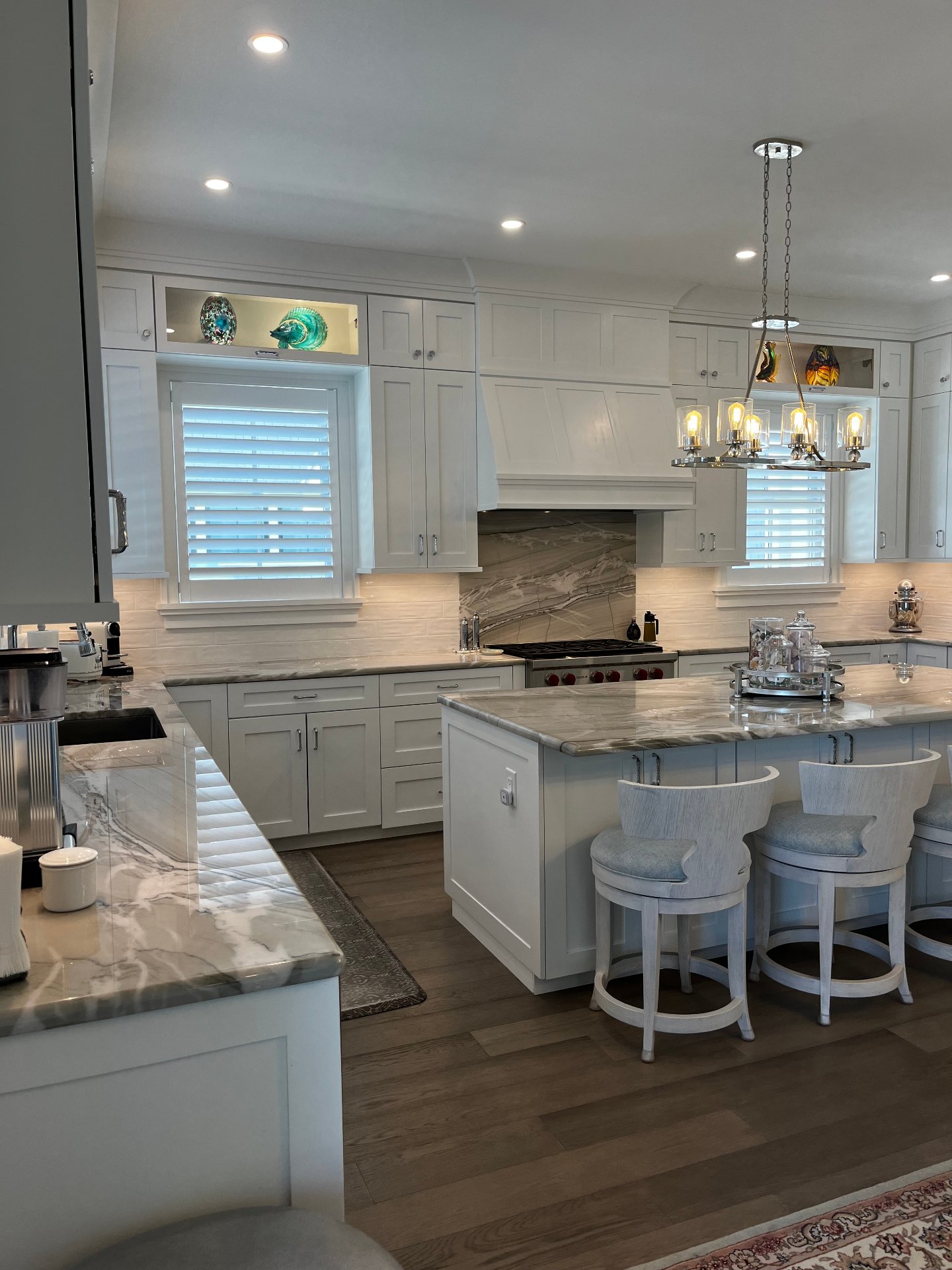 White shaker kitchen with custom hood over a quartzite slab backsplash, lit display cabinets, and a pendant light over the island